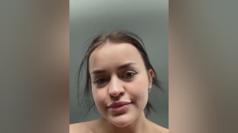 Media: Video of a young woman with light brown skin, brown hair, and dark eyes, wearing minimal makeup, set against a blurred gray background.