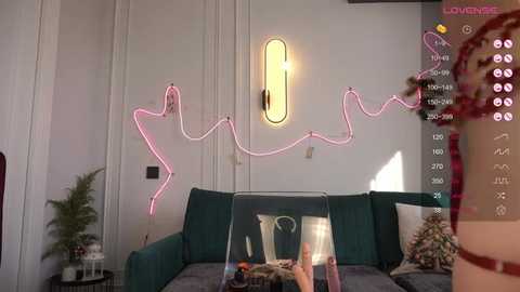 Media: Video of a modern living room with a pink neon sign spelling \"SAY\" above a teal sofa, featuring a hand holding a phone displaying a video call.