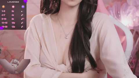 Media: Video of a young East Asian woman with long black hair, wearing a low-cut white lace bra and robe, seated against a soft pink backdrop.