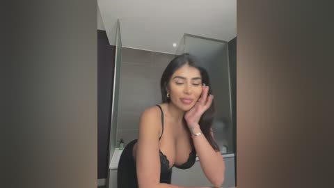 Media: A video of a young woman with long, dark hair, wearing a black lace bra, leaning on her elbows in a modern bathroom with grey tiles and a glass shower door.