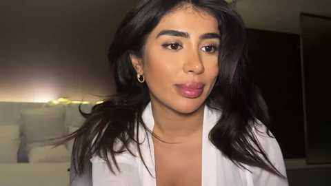 Media: Video of a light-skinned woman with long, dark hair, wearing a white shirt, puckering her lips. Background shows a blurry room with a TV and a bookshelf.