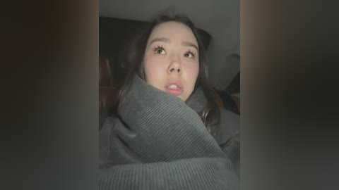 Media: A video of a young Asian woman with light skin, long dark hair, and wide eyes, wearing a grey sweater, looking startled and trapped under a blanket.