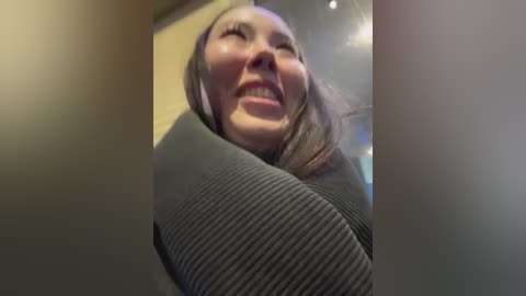 Media: Video of a smiling woman with light skin, brown hair, and wearing a dark gray sweater, captured from a low angle, emphasizing her joyous expression.