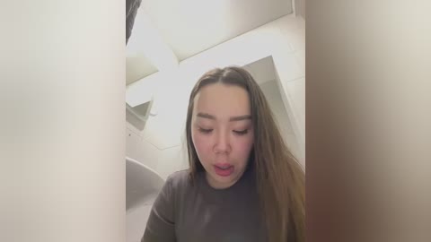 Media: A video captures a young Asian woman with long brown hair and fair skin, wearing a black shirt, making a surprised face. She stands in a dimly lit, narrow hallway with white walls and a sloped ceiling.