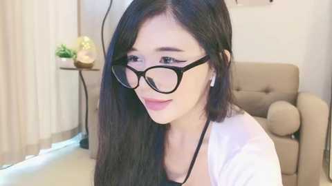 Media: Video of a young East Asian woman with long black hair, wearing black-rimmed glasses, white top, and white headphones, seated indoors near a beige armchair, in a modern, well-lit room with white curtains.