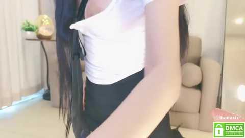 Media: Video of a woman with long black hair, wearing a white blouse and black skirt, standing in a modern living room with beige furniture and potted plants.