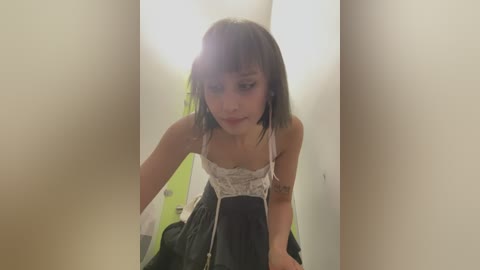 Media: Video of a young woman with shoulder-length brown hair, wearing a white lace camisole and a black skirt, leaning forward, blurred background, indoors.