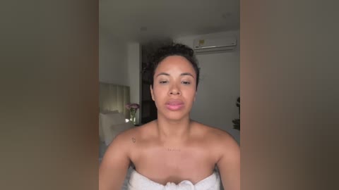 Media: Video of a confident, light-skinned woman with short, curly hair, wearing a white towel, standing in a modern, minimalist bathroom with beige walls, a mirror, and a floral arrangement.