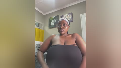 Media: Video of a curvy, dark-skinned woman with glasses, wearing a strapless black dress, standing in a beige-walled room with yellow curtains and framed artwork.