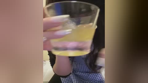 Media: A blurry video of a person holding a glass of orange juice, wearing a black-and-white checkered dress, taken indoors with a dimly lit background.