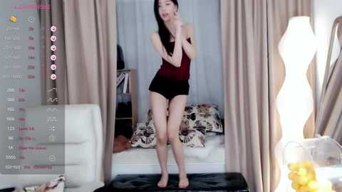 Media: Video of an East Asian woman with long black hair, fair skin, wearing a dark tank top and black shorts, standing barefoot in a cozy, dimly lit living room with a floral-patterned couch, beige curtains, and a tall, modern lamp.