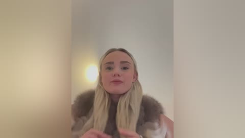 Media: A video of a fair-skinned, blonde woman with straight hair, wearing a fur-trimmed coat, standing in a dimly lit hallway with soft, diffused lighting.