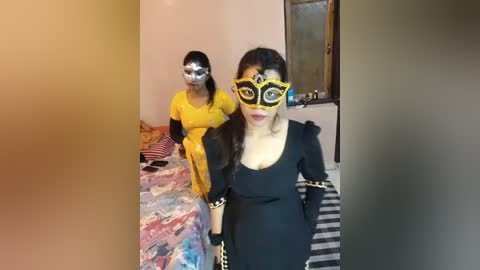 Media: Video of two women in a bedroom: one in a yellow dress with a silver mask, the other in a black dress with a yellow mask, both with long hair, standing near a striped bed.