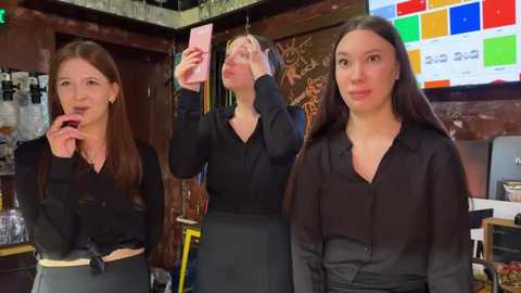 Media: Video of three young women in a cozy, dimly lit bar, wearing black tops and pants. One takes a selfie, another plays with her hair, and the third stands confidently. Background includes colorful bar signs and shelves.