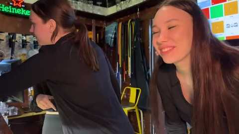 Media: Video of a woman with long brown hair, wearing a black top, smiling in a cozy bar with wooden decor, neon signs, and colorful wall art.