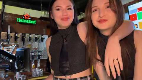 Media: Video of two women in a bar, one with a black top and tie, the other with a tattooed arm, holding hands. Background includes a Heineken sign, bar equipment, and a flat-screen TV.