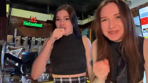 Media: Video of two young women, one with straight black hair, the other with long brown hair, both wearing black crop tops, smiling in a dimly lit bar with Heineken signs and bar equipment in the background.