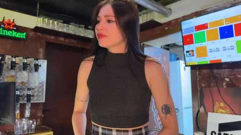 Media: Video of a young woman with long dark hair, wearing a black sleeveless top and plaid skirt, standing in a bar with a colorful menu board and shelves in the background.