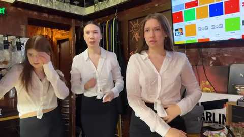 Media: Video of three young women in white shirts and black pants, standing in a dimly lit, cluttered room with a TV displaying colorful squares.