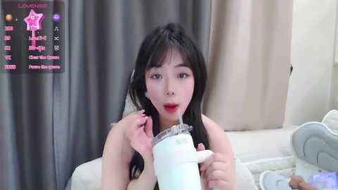 Media: A video of an Asian woman with long black hair, wearing a beige top, drinking from a white cup with a straw, indoors with beige curtains and a bed in the background.