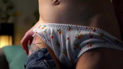 Media: A video of a woman in a ribbed, white tank top with colorful fruit patterns and blue jeans, revealing a tattoo on her left hip. Background includes a blurred lamp and plant.