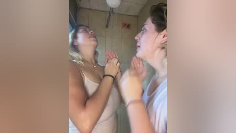 Media: Video of two women in a bathroom, both wearing white tank tops, standing close with hands touching; one has blonde hair, the other dark hair, looking at each other with expressions of joy.