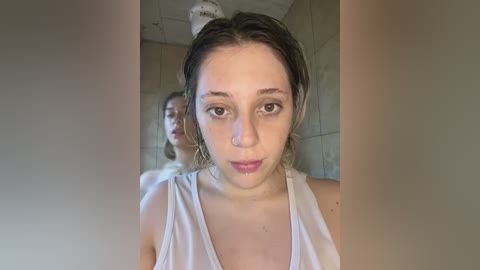 Media: A video of a young woman with fair skin, brown eyes, and short, wet hair, wearing a white tank top, taken from a close-up angle, with a blurred reflection of another person behind her in a dimly lit, tiled bathroom.