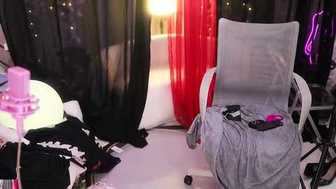 Media: Video of a messy room with a recliner chair, red and black curtains, scattered clothes, pink candle, and a glowing neon sign.