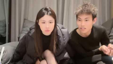 Media: Video of two Asian women sitting side-by-side on a couch. One with long, straight black hair, wears a black puffer jacket, looks introspective. The other with curly, light brown hair, wears a black long-sleeve shirt, appears engaged.