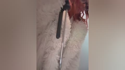 Media: A close-up video of a person with long, curly red hair, wearing a fluffy, white, zip-up jacket, with a black, furry hat. The background is plain and neutral-colored.