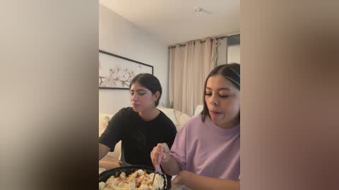 Media: Video of two Latina women with medium skin tones, dark hair, eating from a shared plate on a beige couch, framed by a white wall and a beige curtain.