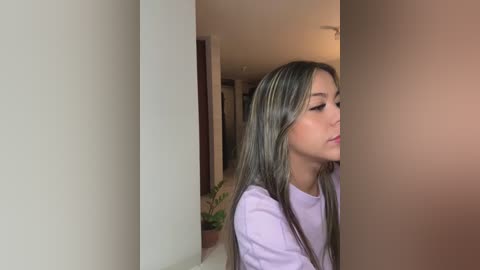 Media: Video of a young Asian woman with long, straight, light brown hair, wearing a light purple shirt, peering through a narrow gap between two white walls in a dimly lit, sparsely furnished room.