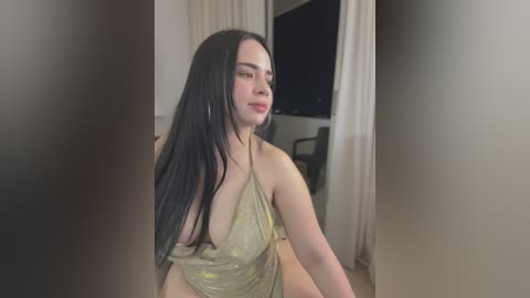 Media: Video of a young woman with long, straight black hair, wearing a gold halter top, sitting indoors near a window. She has a light skin tone and a soft smile.