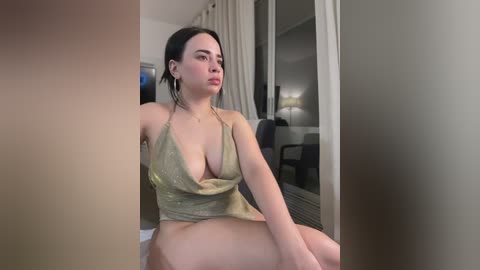 Media: Video of a young woman with fair skin and dark hair, wearing a low-cut olive green top and no pants, sitting indoors by a window.