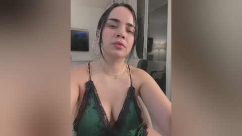 Media: Video of a young woman with dark hair, fair skin, and a medium build, wearing a green lace-trimmed camisole, indoors with blurred background.