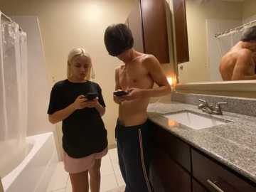 Media: Video of a blonde woman in a black t-shirt and pink shorts, standing beside a shirtless man in dark pants, both holding smartphones in a modern bathroom with a granite counter and mirror.