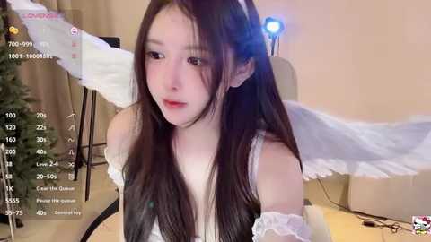 Media: Video of a young East Asian woman with long dark hair, wearing a white off-shoulder dress, sitting at a table with a Christmas tree in the background. She has a neutral expression.