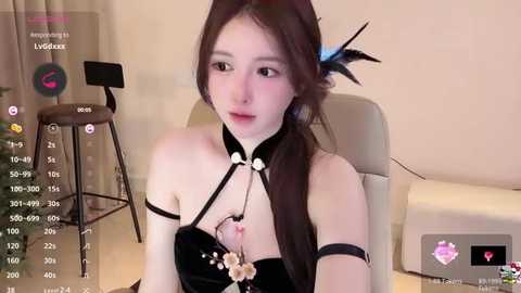 Media: Video of an East Asian woman with fair skin, long brown hair, and a black and white feather accessory. She wears a revealing, black, floral-patterned bodysuit. Background shows a beige wall, a stool, and a chair.