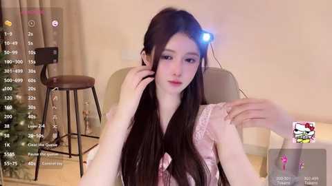 Media: Video of a young Asian woman with long black hair, wearing a pink lace top, seated in a modern beige room with a stool, holding a phone.