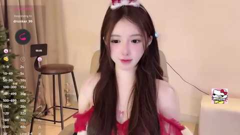 Media: Video of a young Asian woman in a red, off-shoulder Santa dress, with a white Santa hat, sitting in a modern living room with a Christmas tree and stool.