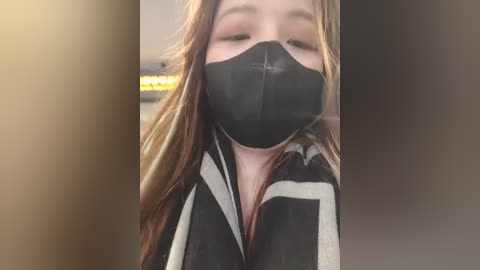 Media: Video of a young woman with fair skin, long brown hair, and a black face mask. She wears a black and white striped shirt. The background is blurred, featuring a yellow sign.