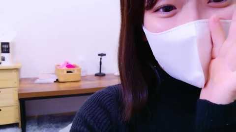 Media: A video of a young Asian woman with long brown hair, wearing a white surgical mask, seated in an office with a wooden desk, pink flowers, and a white box.