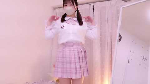 Media: Video of an Asian girl in a white sweater, pink plaid skirt, and face mask, standing in front of a mirror, with a white curtain background.