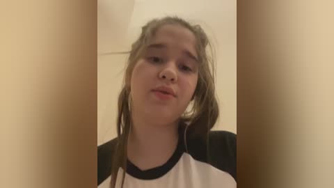 Media: Video of a young girl with light skin, long brown hair, and a pensive expression. She wears a white and black sports shirt indoors against a plain beige wall.