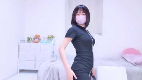 Media: A video of an East Asian woman with straight black hair and light skin, wearing a black, form-fitting dress with a zipper, and a mask, standing in a bright, minimalistic bedroom with white furniture and a bed.