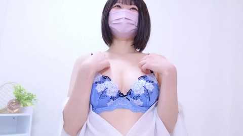 Video of an Asian woman with a bob haircut, wearing a blue lace bra with white floral designs, a face mask, and a white lab coat, standing against a white background.