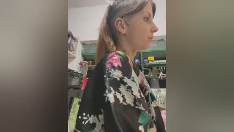 Media: A video of a woman with light skin, brown hair, wearing a black kimono with floral patterns, standing in a cluttered room with shelves and various items in the background.