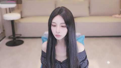 Media: Video of a young Asian woman with long black hair, wearing an off-shoulder striped top, sitting on a blue carpet in a modern living room with beige couches and a round stool.