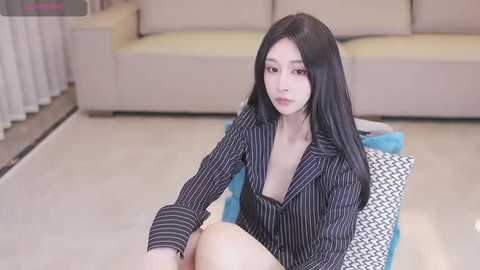 Media: Video of a young Asian woman with long black hair, wearing a black pinstriped blazer, sitting on a blue cushion in a modern living room with beige couches and white curtains.