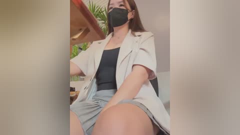 Media: Video of a woman with a light complexion, wearing a black face mask, beige lab coat, black top, and grey shorts, sitting in a room with a wooden desk and green plants.